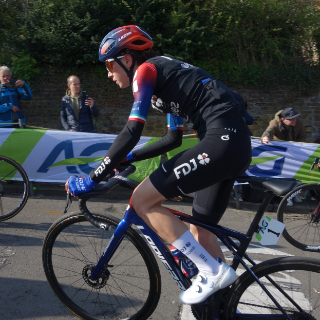 Die professionelle Radsportlerin Katie Archibald fährt mit Helm, Schutzbrille, Handschuhen und Schuhen ein Fahrrad auf einer Straße neben einer Menge Menschen. Im Hintergrund gibt es ein Banner mit Text, eine Wand, Bäume und ein Gebäude.