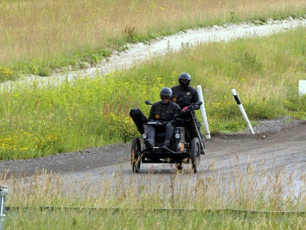 Zwei Polizisten in schwarzen Uniformen und Helmen fahren auf einem dreirädrigen Motorrad über einen mit Gras und Pflanzen gesäumten Schotterweg, mit weißen Pfosten im Hintergrund.
