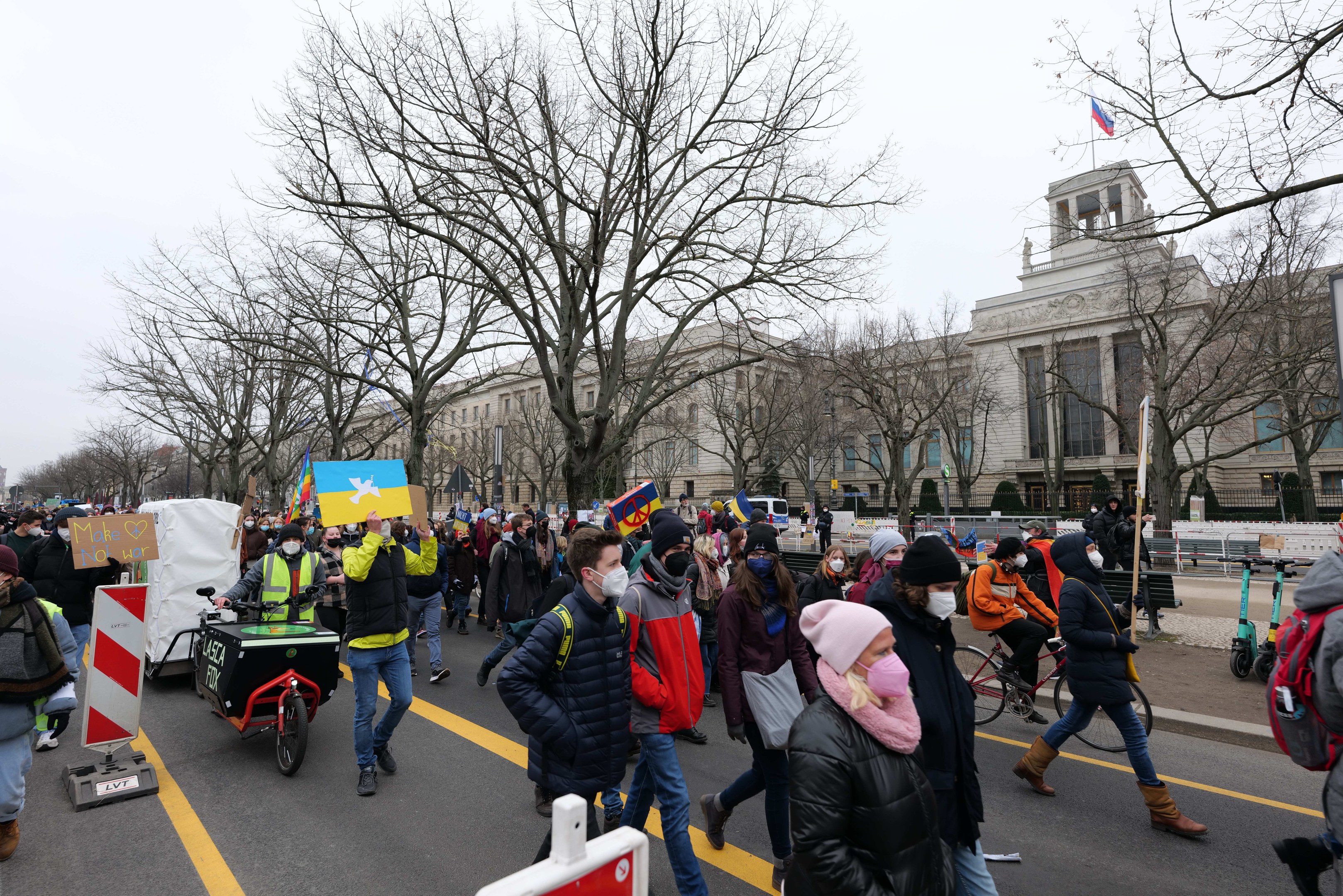 Eine große Gruppe von Menschen marschiert auf einer Demonstration in Washington, D.C. und hält Schilder und fährt Fahrräder, mit Bäumen und einem klaren blauen Himmel im Hintergrund.