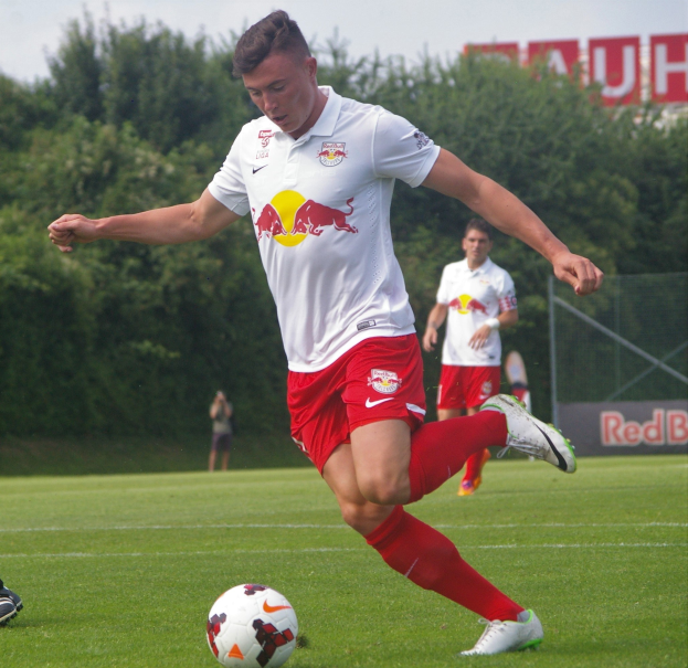 Ein Profifußballer kickt einen Ball auf einem Rasenfeld mit Bäumen und einem klaren blauen Himmel im Hintergrund, mit "RB Leipzig" auf einer Tafel dahinter.