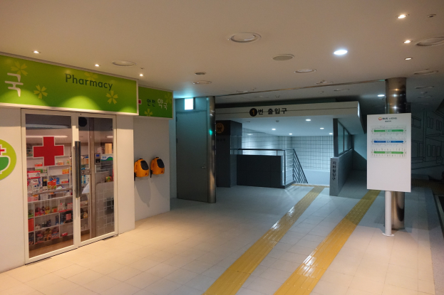 Apotheken-Eingang mit einem "Apotheke"-Schild auf der linken Seite, einer Tafel mit Text auf der rechten Seite, beleuchteten Lampen darüber und Hintergrundobjekten.