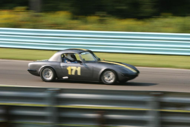 Ein Auto mit einem Helmträger fährt eine Straße entlang, die von Barrieren gesäumt ist, mit Bäumen im Hintergrund.