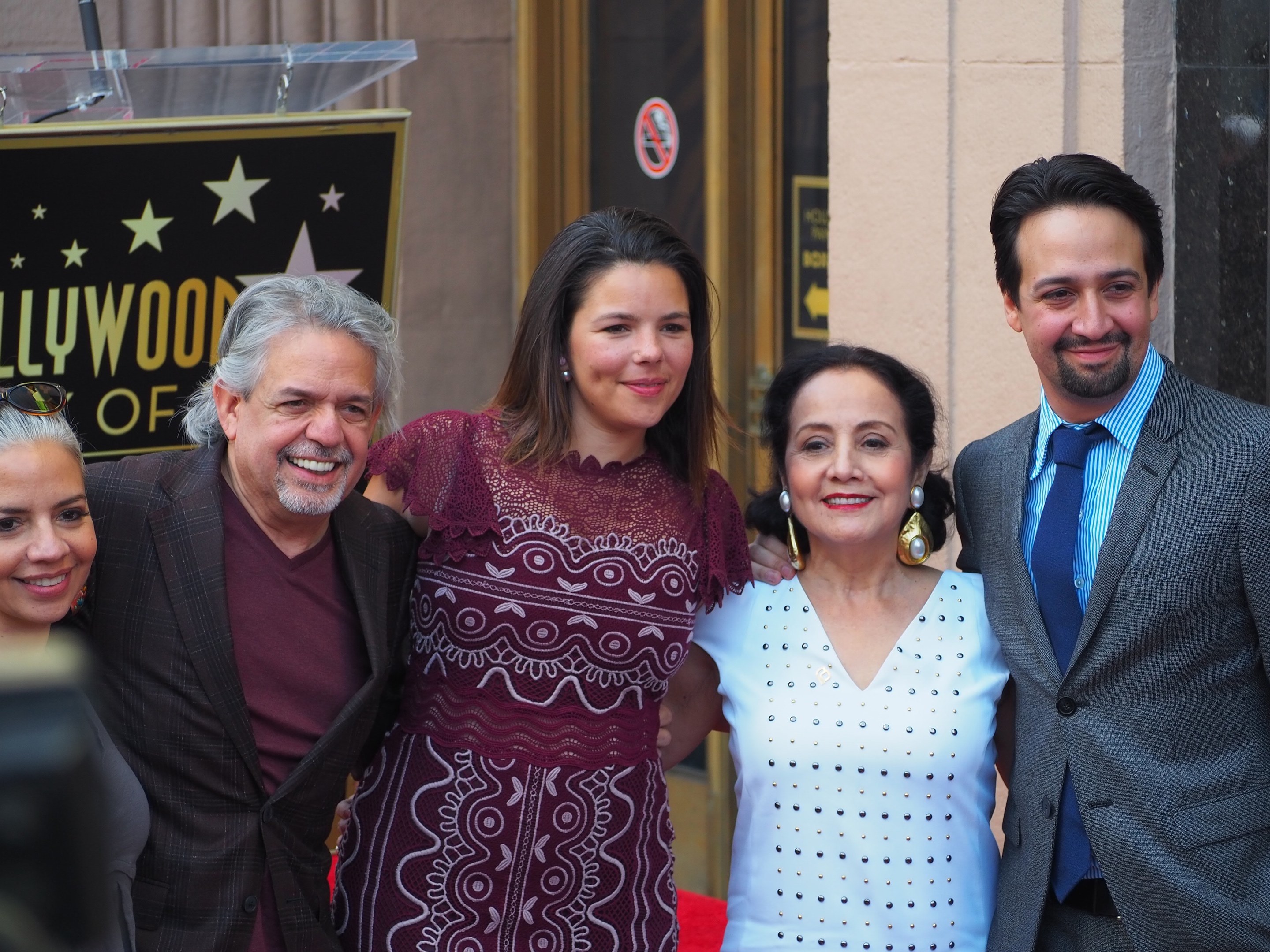 Gruppe von Menschen auf einem roten Teppich bei der Zeremonie des Hollywood Walk of Fame, mit einer Kamera auf der linken Seite, einer Tafel mit Text, einem Glastisch und einer Wand im Hintergrund.