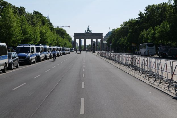Lange Reihe von Polizeiwagen auf der Straße vor dem Reichstagsgebäude in Berlin, Deutschland, mit Fahrradfahrern und Fußgängern, Barrieren, Bäumen und einem Bogen mit Statuen im Hintergrund.