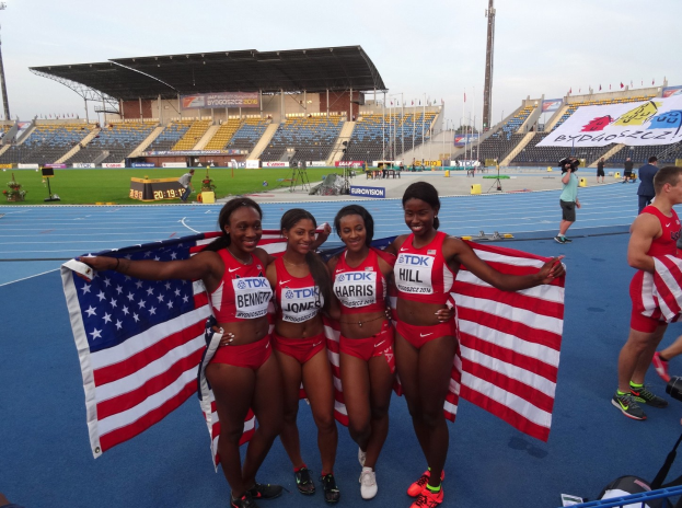Vier Frauen in Sportbekleidung stehen auf einer Laufbahn, halten amerikanische Flaggen und strahlen, mit Zuschauern und Stadionelementen im Hintergrund, feiern ihren 4x400m-Staffelsieg.