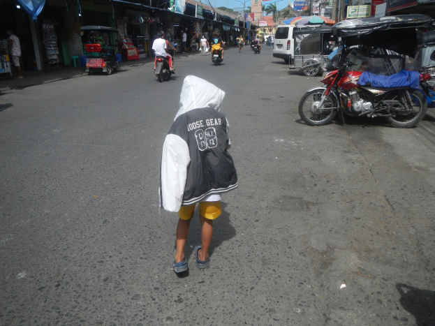 Ein junger Junge geht eine Straße entlang mit Motorrädern, anderen Fahrzeugen und Gebäuden auf beiden Seiten unter einem klaren blauen Himmel.