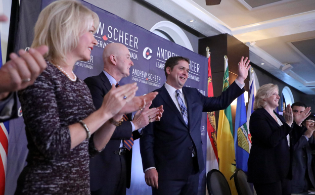 Eine Gruppe von Menschen, die vor Fahnen stehen und jubeln, mit einem Tisch mit Gegenständen rechts daneben und Stühlen, einem Banner und einer Wand im Hintergrund, unter einem Dach mit Lichtern und einem Ventilator.