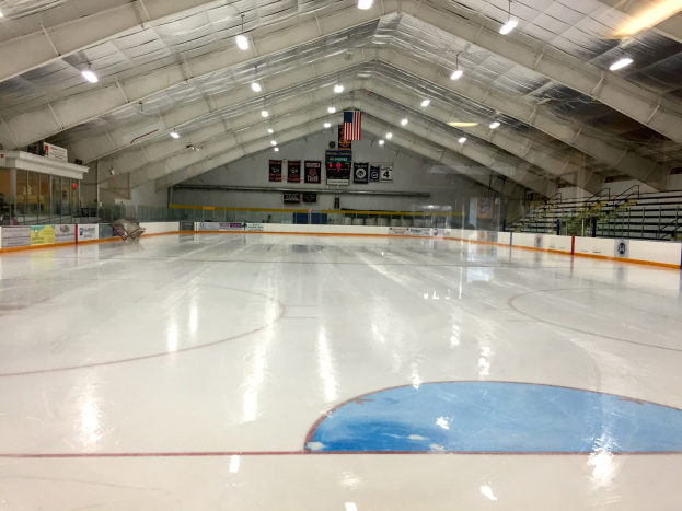 Große Indoor-Eisfläche mit Deckenbeleuchtung, Werbetafeln, Treppen mit Geländern und wenigen Menschen in einer Sportanlage.