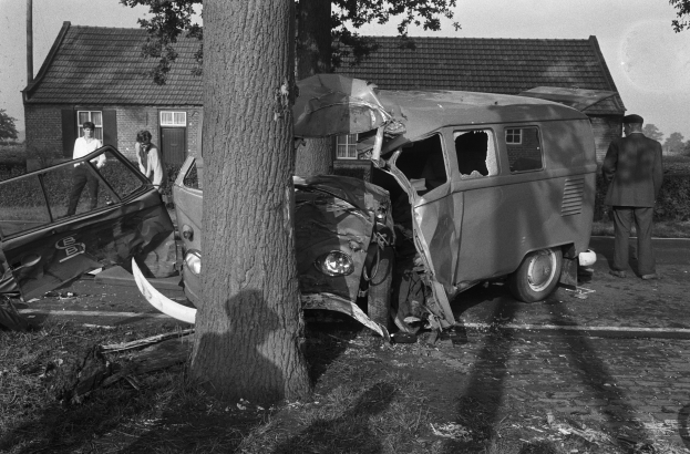 Ein Schwarz-Weiß-Bild eines verunglückten Autos am Straßenrand, mit einem Baum im Vordergrund, einigen Menschen in der Nähe und Häusern, Bäumen und einem Mast im Hintergrund.