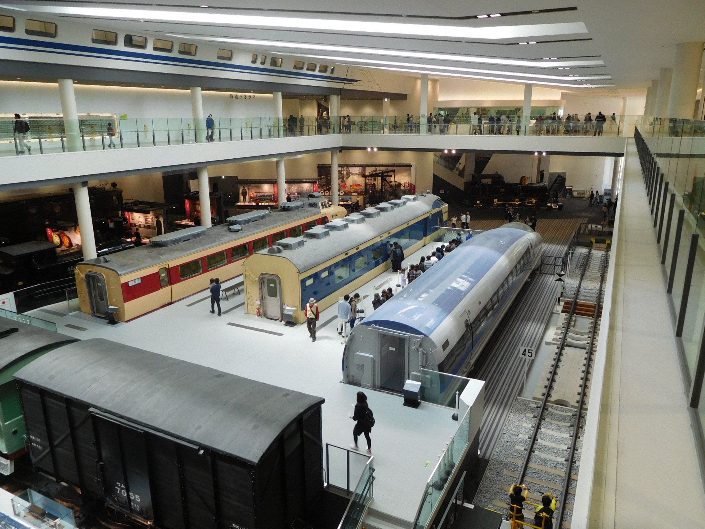 Ein großer Museumsraum mit verschiedenen Bahnen in unterschiedlichen Größen und Farben, bei dem Menschen die Ausstellungsstücke und architektonische Details wie Geländer, Säulen und Deckenleuchten betrachten.