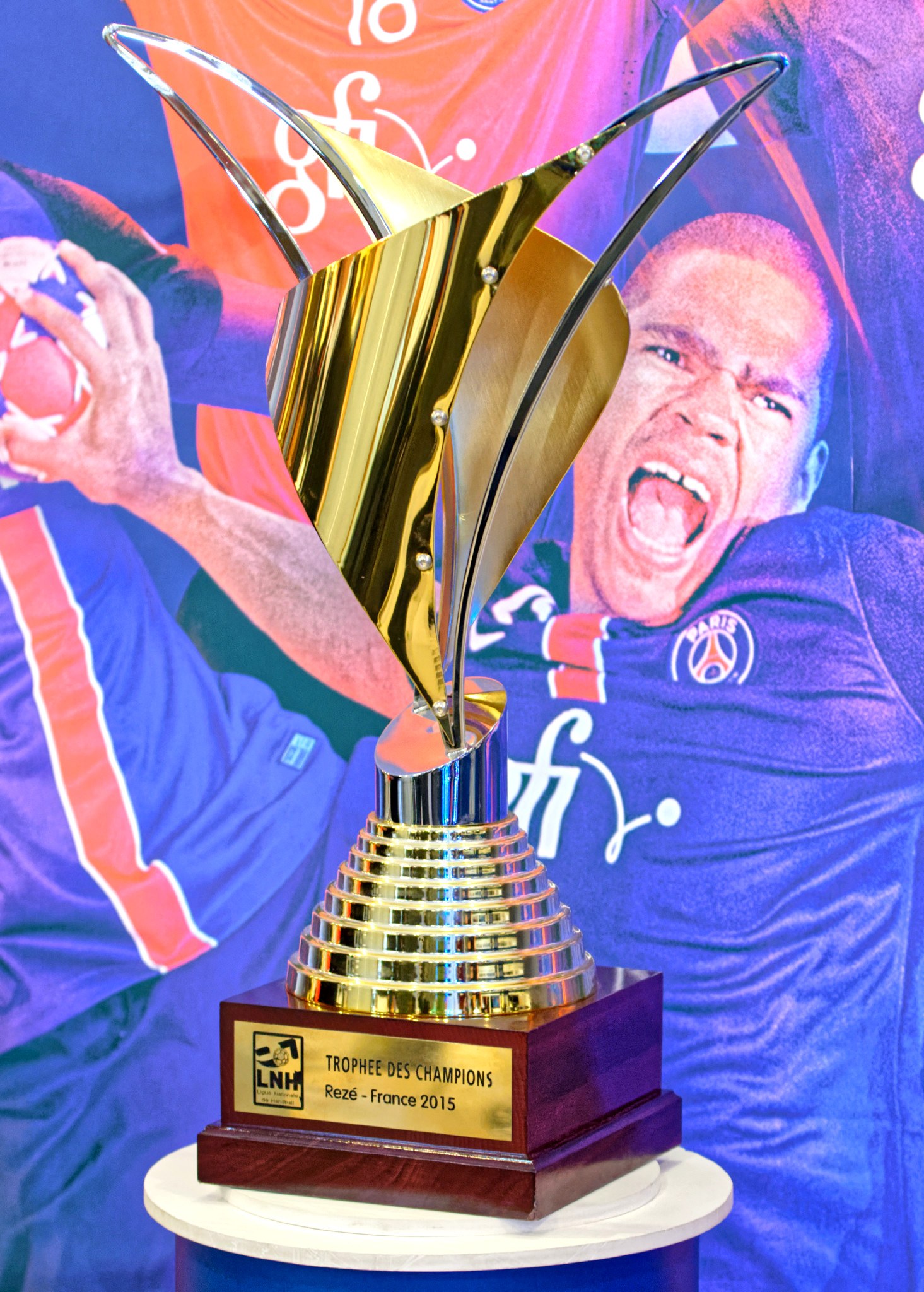 Eine silberne Trophäe auf einem Tisch mit einem unscharfen Banner im Hintergrund, das ein Personenbild zeigt.