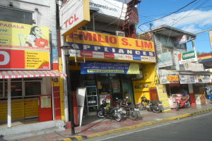 Eine belebte Straßenszene mit Gebäuden, geparkten Motorrädern, Straßenmasten, Straßenlaternen, Strommasten, Stromkabeln, Bäumen und einem bewölkten Himmel, mit einem Geschäft namens "Wilio S Lim Elektrogeräte" im Vordergrund.
