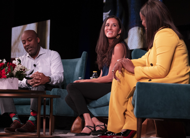 Drei Personen sitzen auf einer Couch vor einem Bildschirm, mit einer Vase und einer Flasche auf dem Tisch dazwischen, während einer Podiumsdiskussion über Geschlechtergerechtigkeit und Frauenrechte.