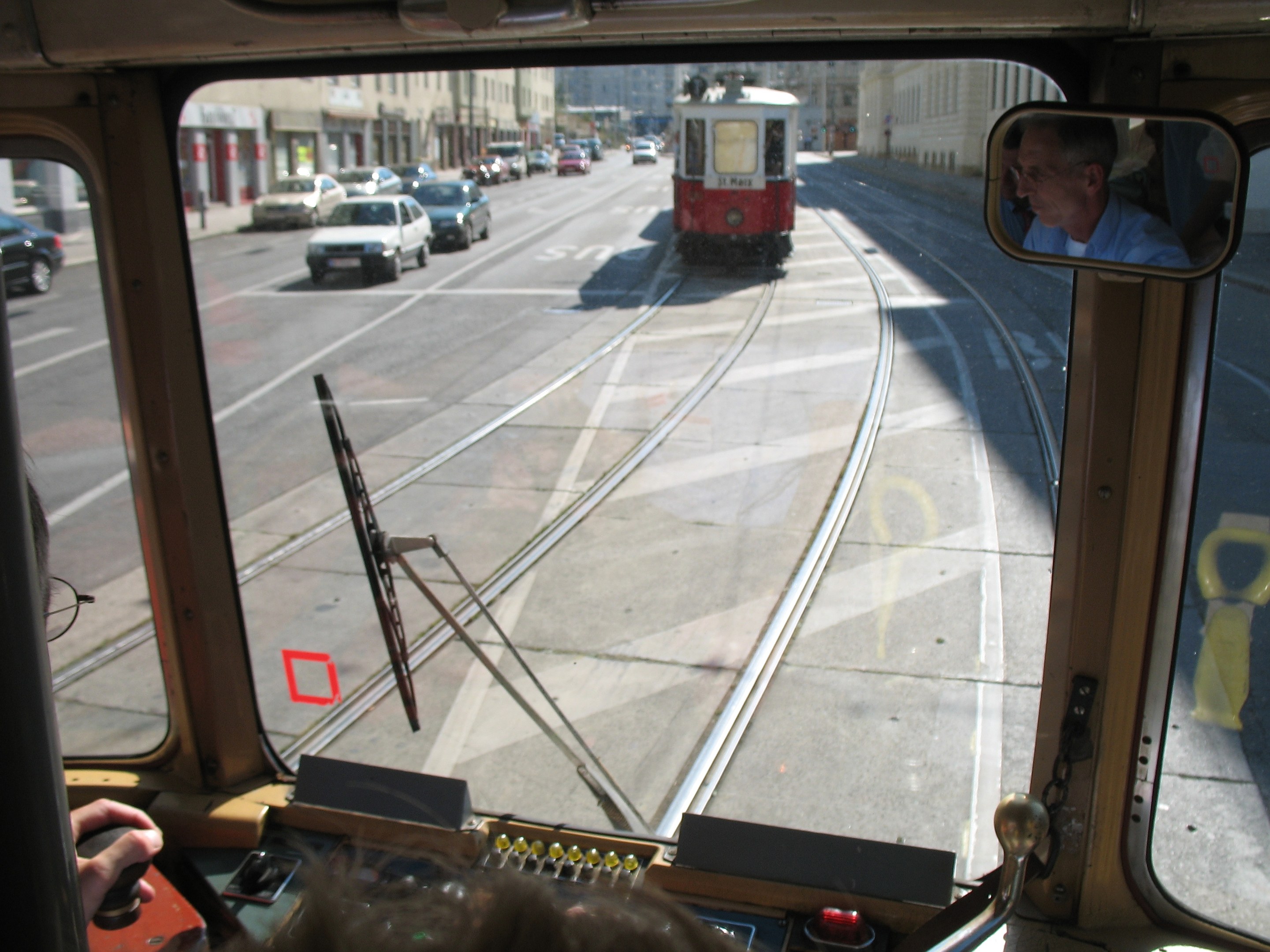 Ein Mann fährt eine Straßenbahn auf einer Stadtstraße mit anderen Fahrzeugen, Gebäuden und einem klaren blauen Himmel im Hintergrund.