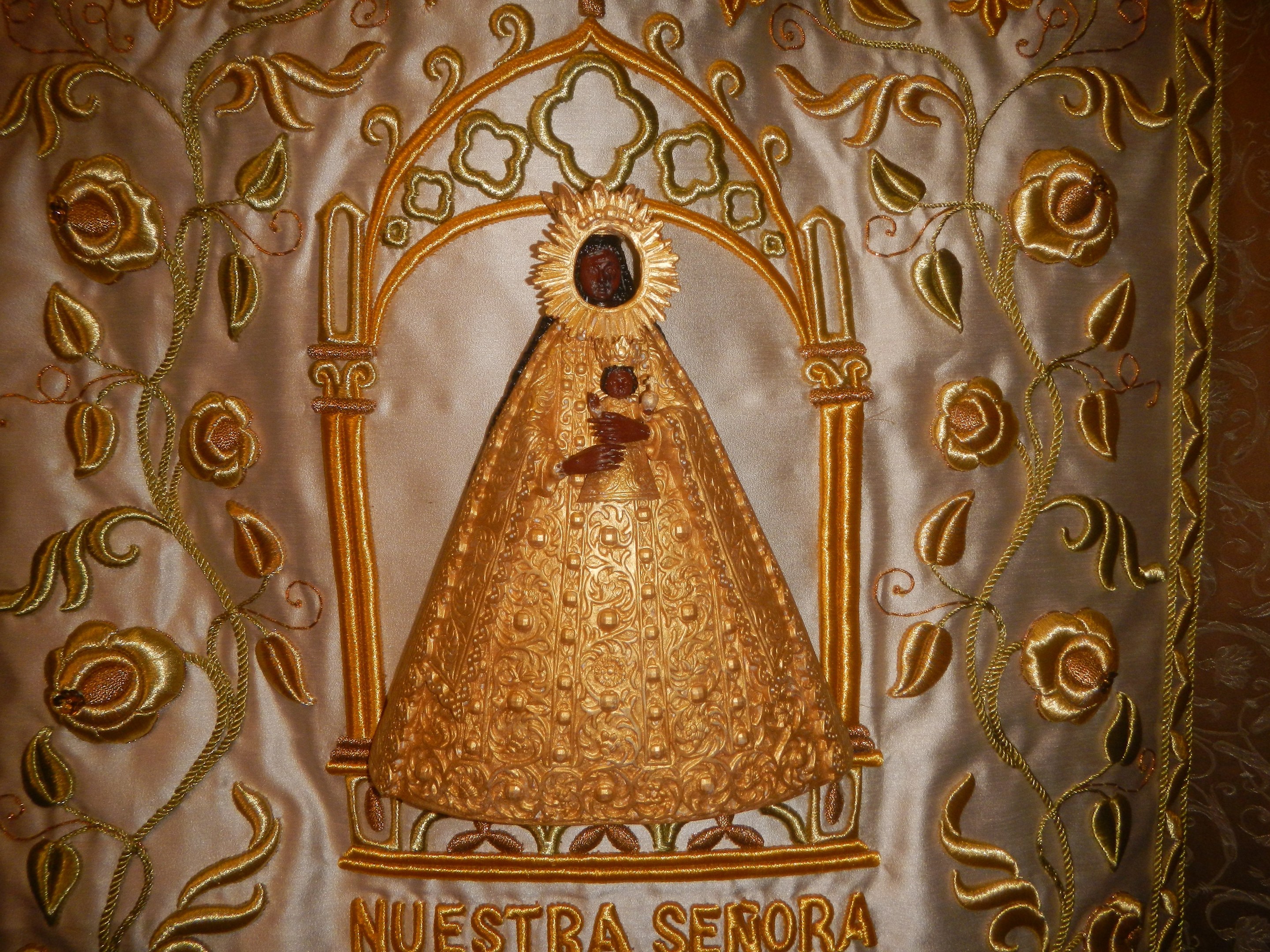 Goldene Stickerei mit einer seren Frau in einem hochgeschlossenen, langärmligen Kleid mit einem Dutt, umgeben von detaillierten Designs und prächtigen Texten in einem goldenen Rahmen.