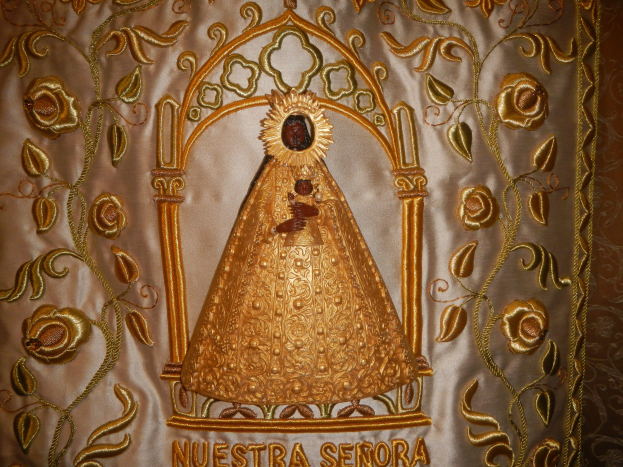 Goldene Stickerei mit einer seren Frau in einem hochgeschlossenen, langärmligen Kleid mit einem Dutt, umgeben von detaillierten Designs und prächtigen Texten in einem goldenen Rahmen.