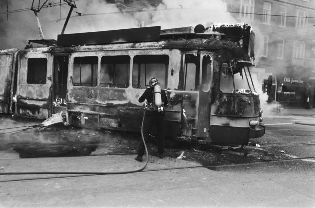 Schwarzes Bild eines Mannes in einem Helm, der eine Pfeife h├Ąlt und neben einer ausgebrannten Stra├čenbahn mit aufsteigendem Rauch steht, vor einem Geb├Ąude mit Fenstern und Dr├Ąhten.