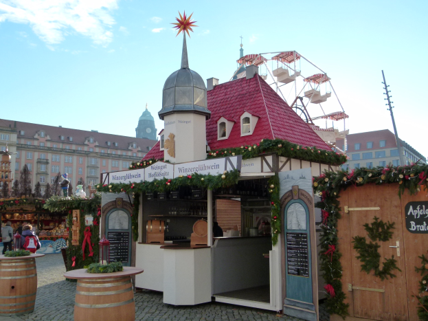 Ein kleines Gebäude mit rotem Dach und einem Riesenrad davor, umgeben von Festdekorationen und Menschen, mit Gebäuden, Bäumen und einem bewölkten Himmel im Hintergrund auf einem Oktoberfest in München, Deutschland.