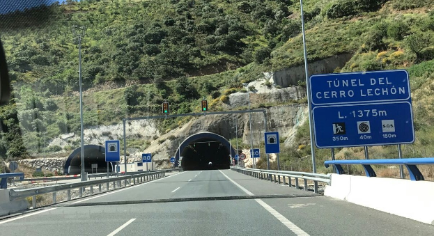 Auto fährt durch einen Tunnel mit Geländern, Verkehrszeichen, Pfosten, Ampeln, Bäumen, Felsen und einem klaren blauen Himmel.