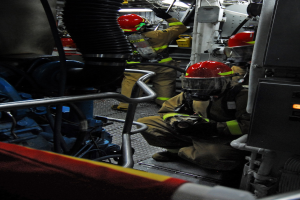 Feuerwehrleute in Helmen und Schutzausrüstung arbeiten an einem Feuerlöschwagen mit Rohren und Gegenständen im Hintergrund, unter einem Licht oben.