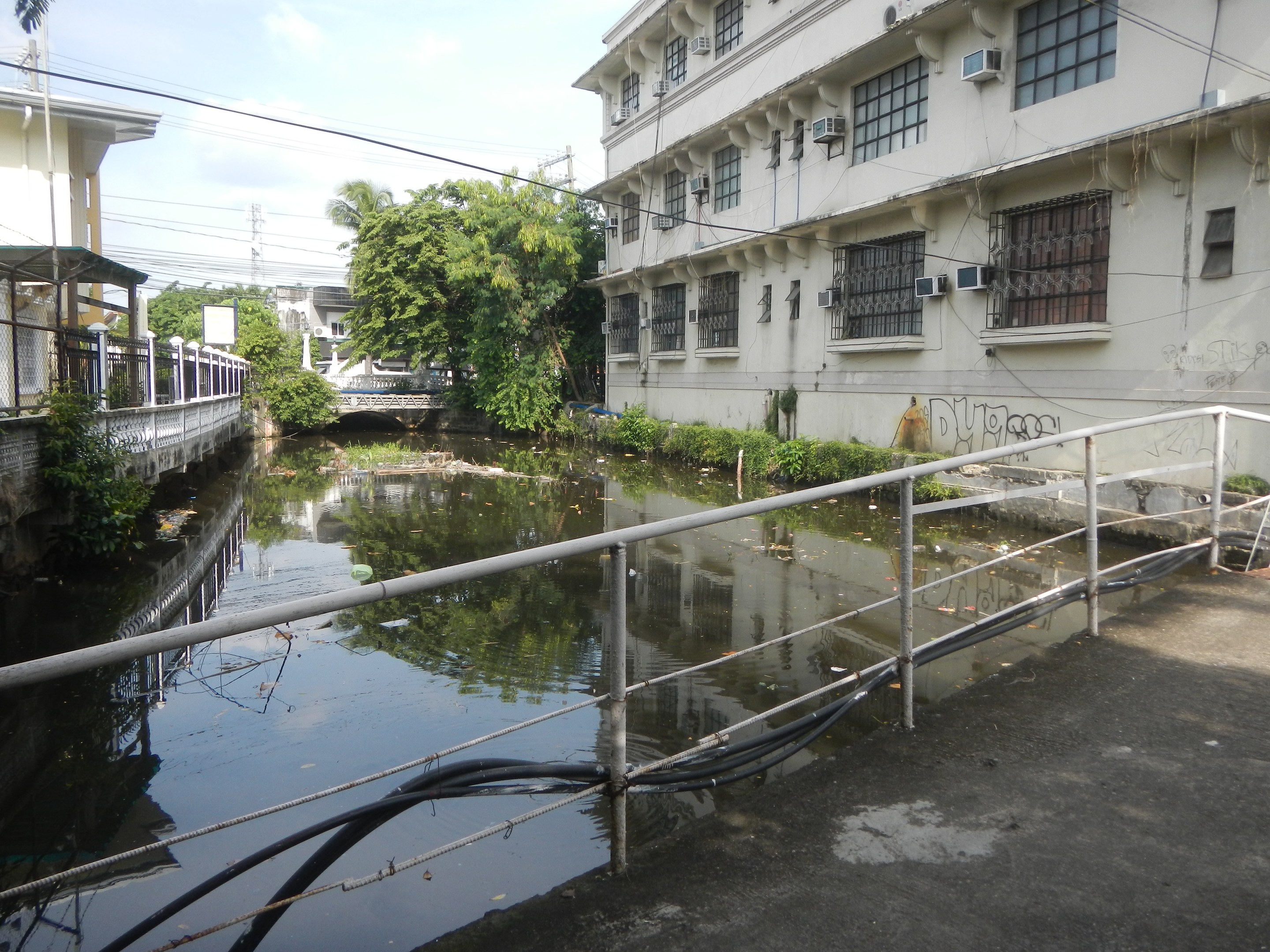 Ein kleiner Kanal verläuft durch eine Stadtstraße, flankiert von hohen Gebäuden, mit Wasser, Geländern, Rohren, Pflanzen, Bäumen, Strommasten mit Kabeln und einem bewölkten Himmel im Hintergrund.