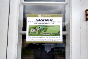 Glasstür mit einem Schild "Geschlossene öffentliche Toiletten in der Nähe des Hauptparkplatzs" und einem unklaren Objekt auf der rechten Seite.