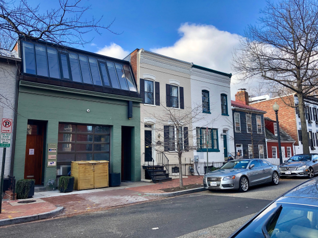 Eine Straße in einer Nachbarschaft in Philadelphia, gesäumt von geparkten Autos, Gebäuden, Bäumen, Laternen, Schildern und Holzkisten unter einem bewölkten Himmel.
