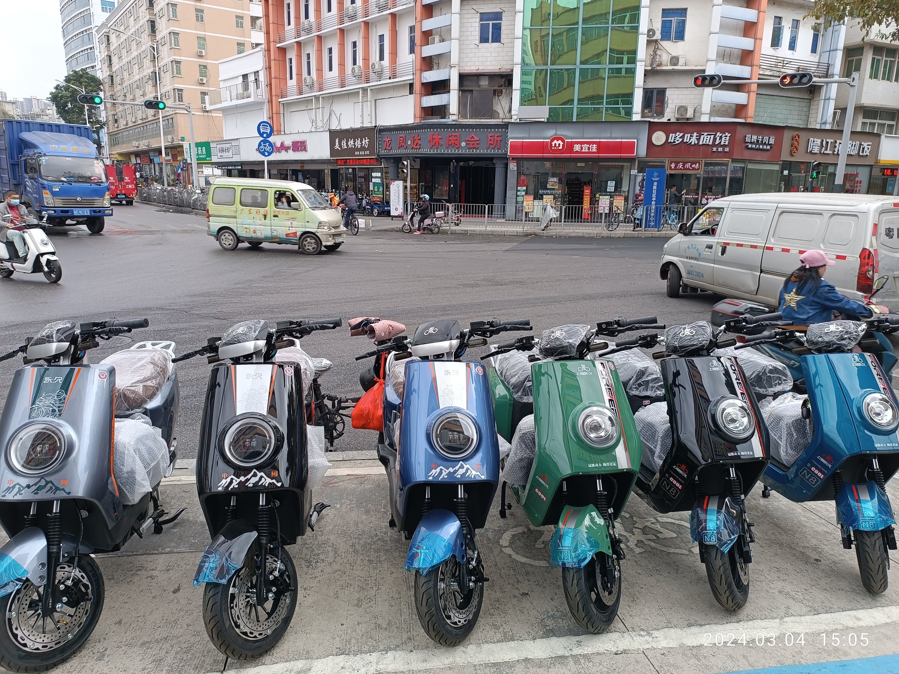 Eine Reihe von Elektro-Roller, die an einer Straße geparkt sind, mit einer Person mit einer Kappe davor, vor einem Hintergrund von Gebäuden, Ampeln, Bäumen und einem klaren Himmel.