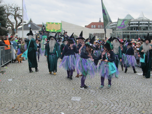 Eine Gruppe von Menschen in Hexenkostümen, die schwarze und lila Kleider und Hüte tragen und die einige Fahnen halten, gehen eine Kopfsteinpflasterstraße entlang, neben einem Geländer mit Gebäuden, Bäumen und einem klaren blauen Himmel im Hintergrund.