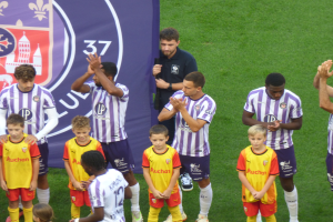 Eine Gruppe junger Jungs steht auf einem Fußballfeld, klatscht in die Hände, mit einem Banner und Logos im Hintergrund und grünem Gras auf dem Boden.