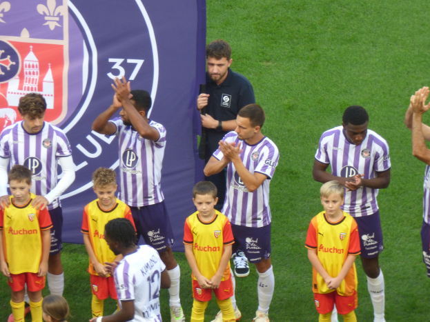Eine Gruppe junger Jungs steht auf einem Fußballfeld, klatscht in die Hände, mit einem Banner und Logos im Hintergrund und grünem Gras auf dem Boden.