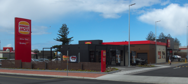 Eine belebte Stadtstraße mit einer Hungry Jacks-Filiale, umgeben von Gebäuden, Bäumen, Straßenlaternen, Lichtern, Fahrzeugen, Fußgängern, Werbetafeln und Grills unter einem bewölkten Himmel.