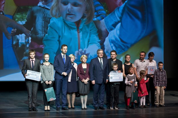 Gruppe von Menschen auf einer Bühne mit Taschen und Urkunden, im Hintergrund ein Bildschirm mit einem Bild eines Kindes und anderen Personen.