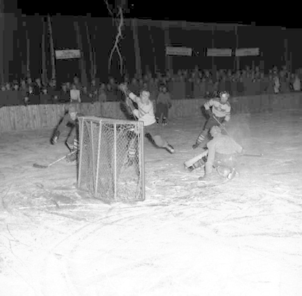 Schwarzes und weißes Bild eines Hockey-Spiels auf einem Eisstadion, mit Spielern in der Nähe eines Tors und Zuschauern im Hintergrund.