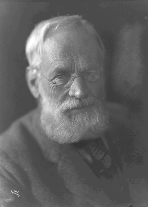 Ein ernst aussehender älterer Mann mit Vollbart und Brille, der eine dunkle Hintergrund hat und in die Ferne schaut.