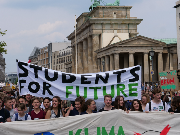 Gruppe von Schülern marschiert in Berlin mit einer bunten "Students for Future"-Schleife vor Gebäuden, Bäumen und Himmel.