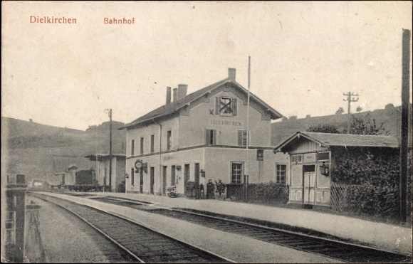 Schwarze und weiße Fotografie eines Bahnhofs in Dielkirchen, Deutschland, mit einem Zug auf den Gleisen, umgeben von Häusern, Strommasten, Bäumen, Menschen und Hügeln im Hintergrund unter einem klaren Himmel.