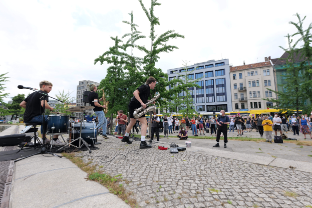 Eine Gruppe von Menschen, die auf einem Bürgersteig Musik machen, mit verstreuter Ausrüstung, vor einem Publikum, mit Bäumen, Gebäuden und einem bewölkten Himmel im Hintergrund.