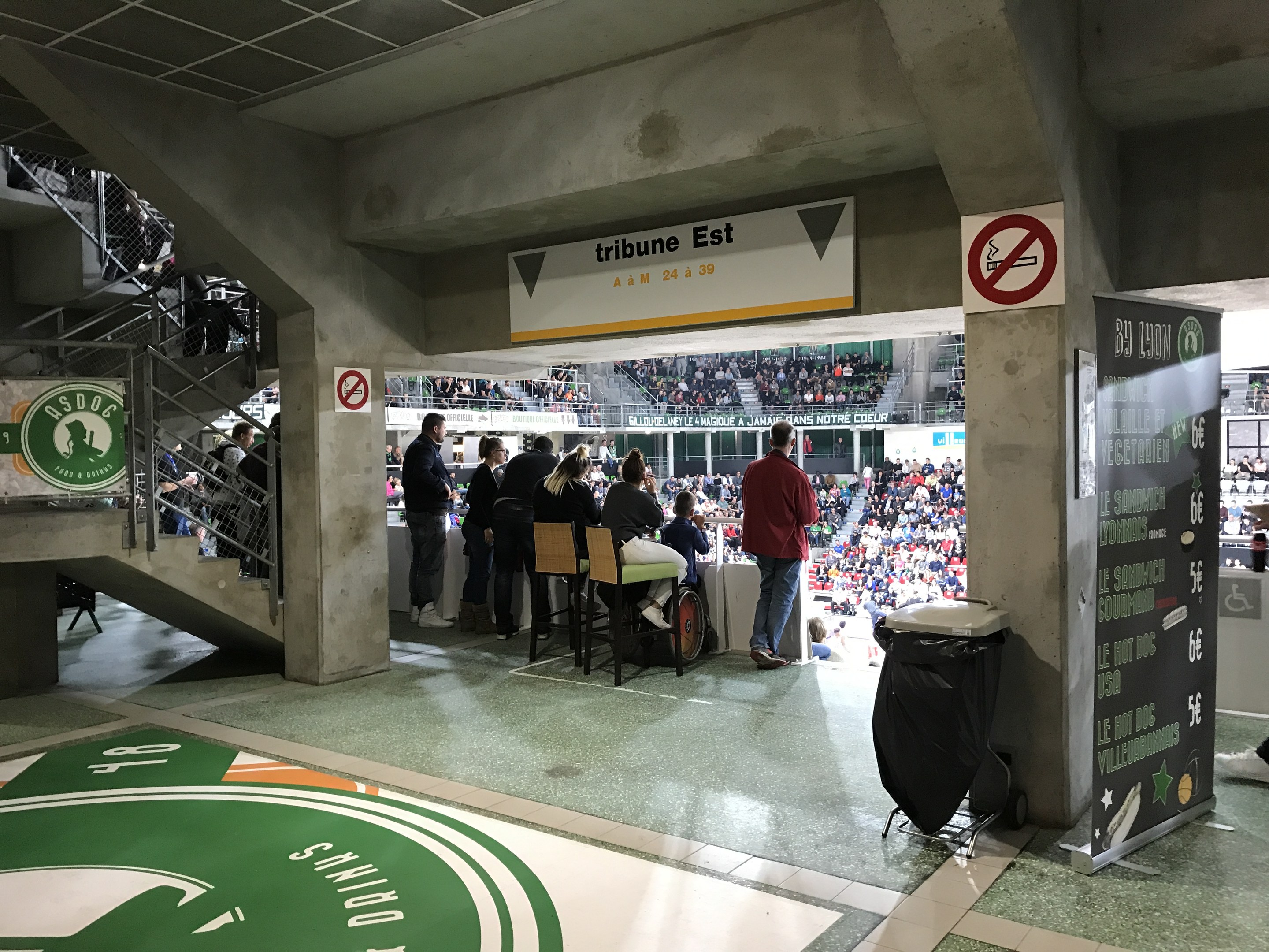 Eine Gruppe von Menschen steht vor einem sitzenden Publikum in einem Stadion mit Schildern, die "Boston Celtics vs New York Jets" lesen, einem Müllcontainer, einer Tafel mit Text, einer Treppe mit Geländern und einer Decke mit Deckenleuchten.