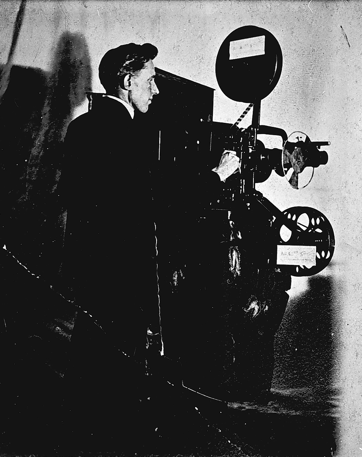 Ein Mann in einem Anzug steht neben einem alten Filmprojektor mit einem ernsten Gesichtsausdruck.