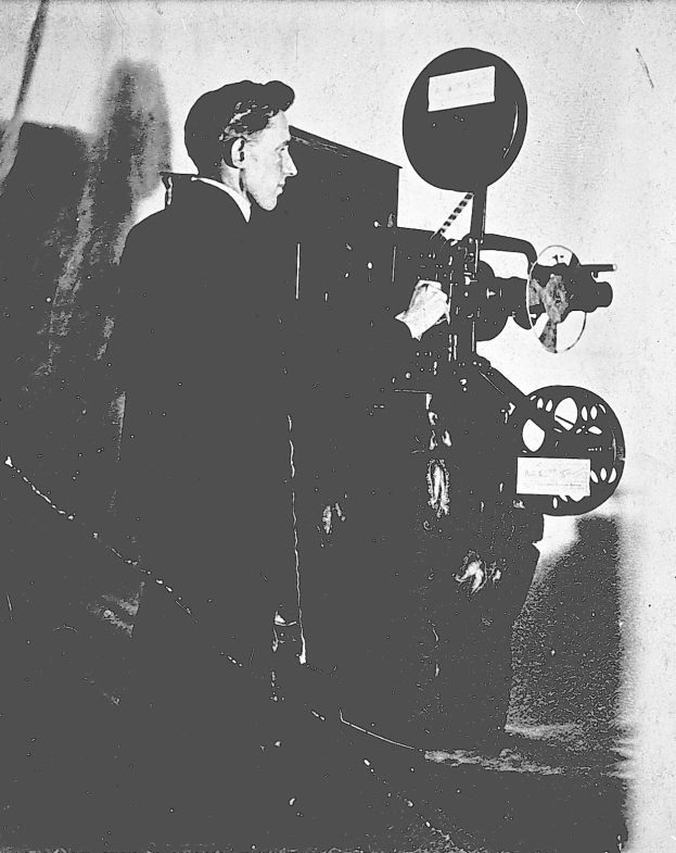 Ein Mann in einem Anzug steht neben einem alten Filmprojektor mit einem ernsten Gesichtsausdruck.