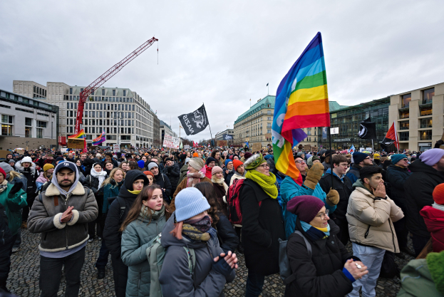 Eine große Gruppe von Menschen mit Fahnen und Spruchbändern vor einem Gebäude während einer LGBTQ+-Rechtsdemo in Berlin, mit Gebäuden, einem Kran und einem bewölkten Himmel im Hintergrund.