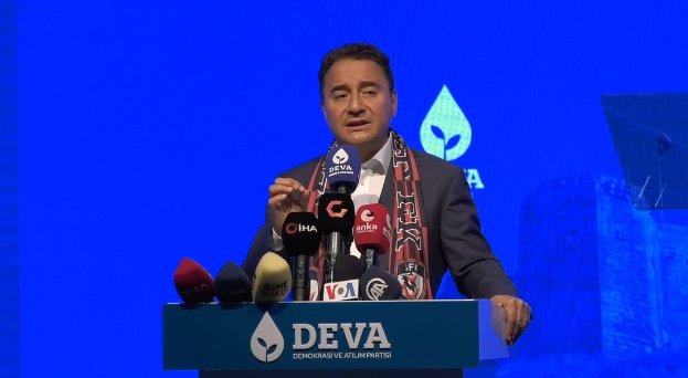 Ein Mann in Mantel und Schal steht an einem Podium mit Mikrofonen und spricht hinter einer Tafel mit dem Text "deva-Geschäftsführer sagt, es sei keine gute Idee für die Zukunft", mit einem Bildschirm und einem Gegenstand im Hintergrund.