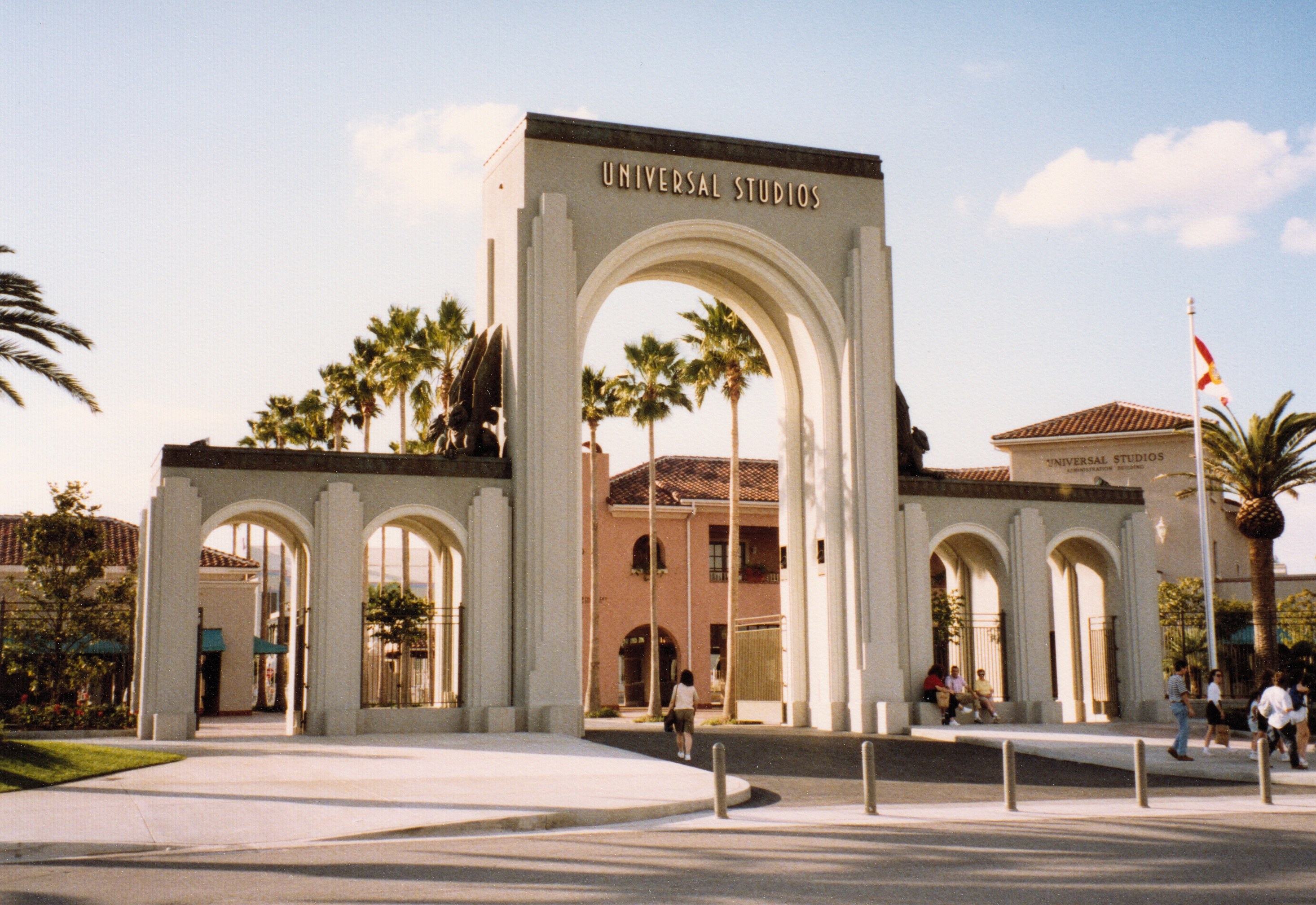Eingang zum Universal Studios Hollywood mit einem Bogen, einer Statue, Menschen, Bäumen, Gebäuden, einer Fahnenstange mit einer Fahne und einem bewölkten Himmel.
