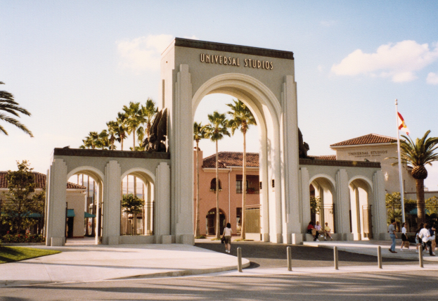 Eingang zum Universal Studios Hollywood mit einem Bogen, einer Statue, Menschen, Bäumen, Gebäuden, einer Fahnenstange mit einer Fahne und einem bewölkten Himmel.