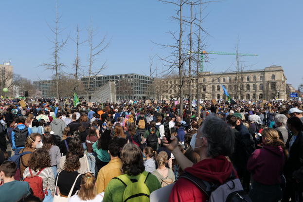 Eine große Menge von Menschen mit Schildern und Tötenschutzwesten vor einem Gebäude mit Fenstern, Bäumen und einem klaren Himmel bei einer Klimaprotest