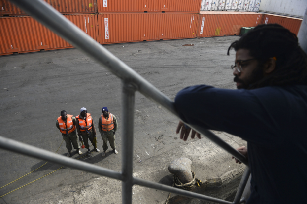 Ein Mann in legerer Kleidung steht auf einem Metallgeländer in der Nähe einer Gruppe von Menschen in orangefarbenen Jacken und Mützen, mit Containern und einer Wand im Hintergrund am Hafen von Los Angeles.