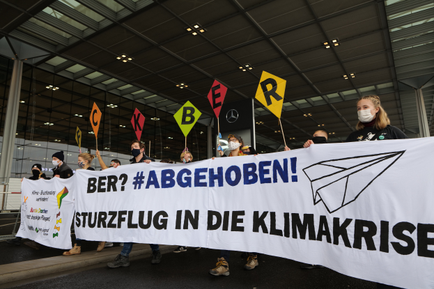 Eine Gruppe maskierter Personen mit einem Banner 'Sturzflug in die Klimakrise' vor einem Glasgebäude mit Säulen, beleuchtet von Deckenlampen.