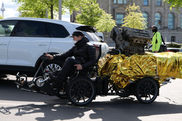 Ein Mann im Rollstuhl mit einem großen Motor daran, umgeben von Fahrzeugen auf einer Straße mit Bäumen, Gebäuden und einem klaren blauen Himmel im Hintergrund, der eine schwarze Jacke und eine Kappe trägt und ein Objekt hält.
