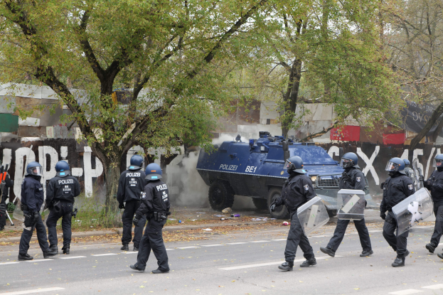 Polizeibeamte in schwarzen Uniformen und Helmen gehen neben einem gepanzerten Fahrzeug eine Straße entlang, einige tragen Schilde, mit Bäumen, Gebäuden und einem klaren blauen Himmel im Hintergrund.